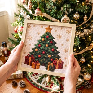 May include: A square framed wall art piece featuring a quilted-style illustration of a green Christmas tree decorated with red and gold ornaments, a gold star topper, and gold snowflakes on a cream background, with three wrapped gifts below.