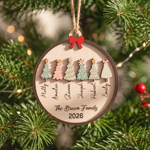 May include: A round wooden Christmas ornament featuring six stylized trees in various colors and patterns. Below each tree is a name: Molly, Amelia, Steven, Joseph, Robert, and Emily. The text The Brown Family 2026 is printed below.