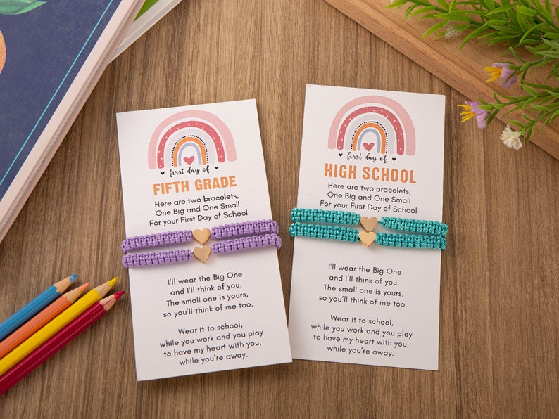 Puede incluir: Dos tarjetas con dise&ntilde;os de arco&iacute;ris y el texto "first day of FIFTH GRADE" y "first day of HIGH SCHOOL". Cada tarjeta tiene dos pulseras con un dije de coraz&oacute;n, una morada y una turquesa.
