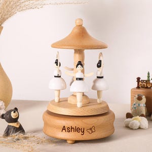 May include: A wooden carousel music box featuring three ballerina figures with white dresses and black hair. The base is made of light-colored wood and is engraved with the name Ashley and a small butterfly icon. The structure has a central pillar.