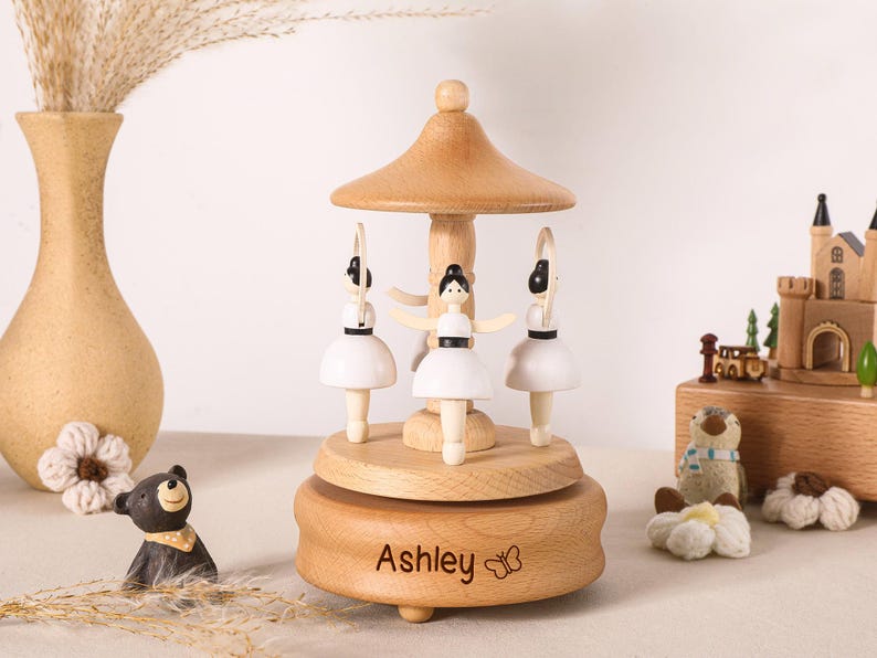 May include: A wooden carousel music box featuring three ballerina figures with white dresses and black hair. The base is made of light-colored wood and is engraved with the name Ashley and a small butterfly icon. The structure has a central pillar.