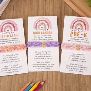 Puede incluir: Tres tarjetas con dise&ntilde;os de arco&iacute;ris y el texto "first day of" seguido de "EIGHTH GRADE", "HIGH SCHOOL" y "PRE-K". Cada tarjeta tiene dos pulseras, una rosa, una morada y una rosa claro, con dijes de coraz&oacute;n dorados. Las tarjetas tienen un mensaje sobre usar las pulseras y pensar el uno en el otro.