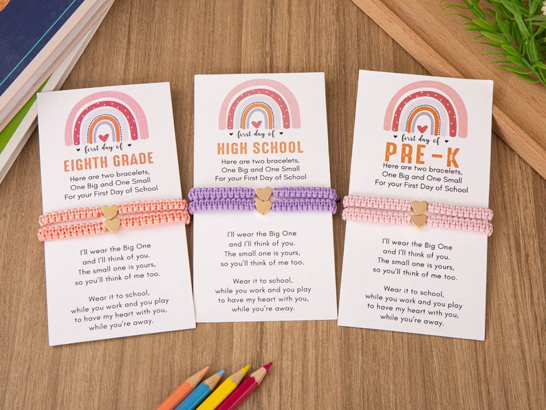 Puede incluir: Tres tarjetas con dise&ntilde;os de arco&iacute;ris y el texto "first day of" seguido de "EIGHTH GRADE", "HIGH SCHOOL" y "PRE-K". Cada tarjeta tiene dos pulseras, una rosa, una morada y una rosa claro, con dijes de coraz&oacute;n dorados. Las tarjetas tienen un mensaje sobre usar las pulseras y pensar el uno en el otro.