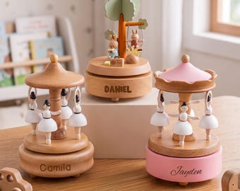 May include: Three wooden music boxes. Two are carousel-style with white-clad ballerinas, one with a natural wood base labeled 'Camila' and one with a pink base labeled 'Jayden'. The center box features a tree, rabbits, and the name 'DANIEL'.