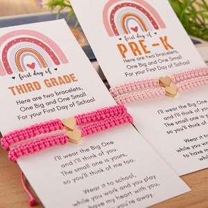 Puede incluir: Dos pulseras de amistad de color rosa con dijes de coraz&oacute;n dorados y una tarjeta con un dise&ntilde;o de arco&iacute;ris y el texto "first day of THIRD GRADE" y "first day of PRE-K". La tarjeta incluye un mensaje sobre llevar las pulseras a la escuela y pensar el uno en el otro.