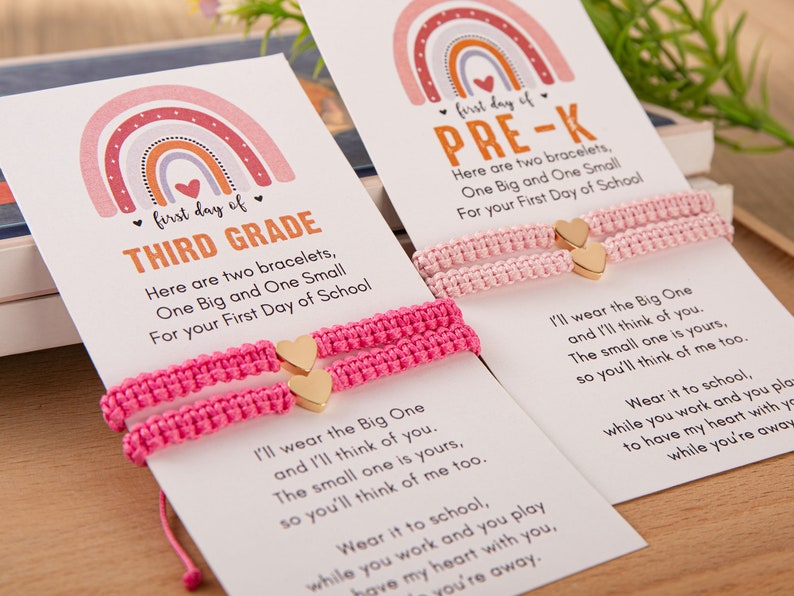 Puede incluir: Dos pulseras de amistad de color rosa con dijes de coraz&oacute;n dorados y una tarjeta con un dise&ntilde;o de arco&iacute;ris y el texto "first day of THIRD GRADE" y "first day of PRE-K". La tarjeta incluye un mensaje sobre llevar las pulseras a la escuela y pensar el uno en el otro.