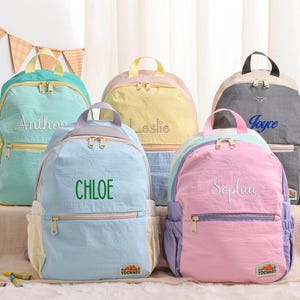 May include: Five colorful backpacks with embroidered names: Anthony, Chloe, Leslie, Joyce, and Sophia. Each backpack features a top handle, front pocket, and side pockets. The backpacks are in shades of blue, yellow, pink, and gray.
