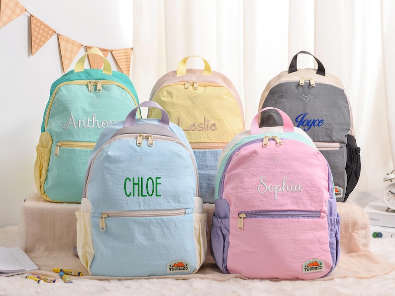 May include: Five colorful backpacks with embroidered names: Anthony, Chloe, Leslie, Joyce, and Sophia. Each backpack features a top handle, front pocket, and side pockets. The backpacks are in shades of blue, yellow, pink, and gray.