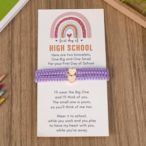 Puede incluir: Una tarjeta con un dise&ntilde;o de arco&iacute;ris y el texto "first day of HIGH SCHOOL" con dos pulseras de amistad moradas con dijes de coraz&oacute;n de madera. La tarjeta dice "Here are two bracelets, One Big and One Small For your First Day of School. I'll wear the Big One and I'll think of you. The small one is yours, so you'll think of me too. Wear it to school, while you work and you play to have my heart with you, while you're away."