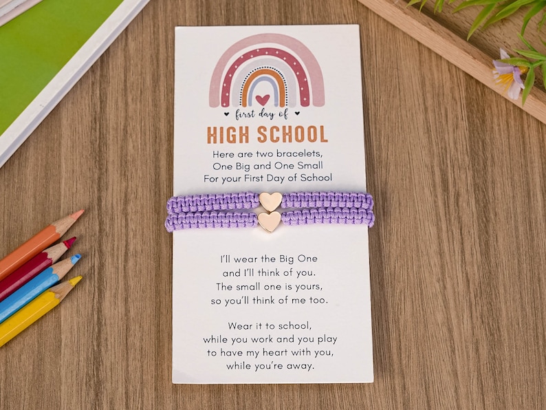 Puede incluir: Una tarjeta con un dise&ntilde;o de arco&iacute;ris y el texto "first day of HIGH SCHOOL" con dos pulseras de amistad moradas con dijes de coraz&oacute;n de madera. La tarjeta dice "Here are two bracelets, One Big and One Small For your First Day of School. I'll wear the Big One and I'll think of you. The small one is yours, so you'll think of me too. Wear it to school, while you work and you play to have my heart with you, while you're away."