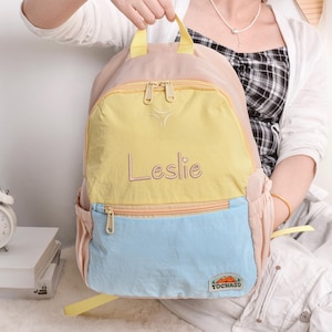 May include: A multi-colored backpack with a light pink body, a yellow front pocket, and a light blue bottom pocket. The name "Leslie" is embroidered in cursive on the yellow pocket. The backpack has a zipper closure and a small logo that says "TOCHASO".