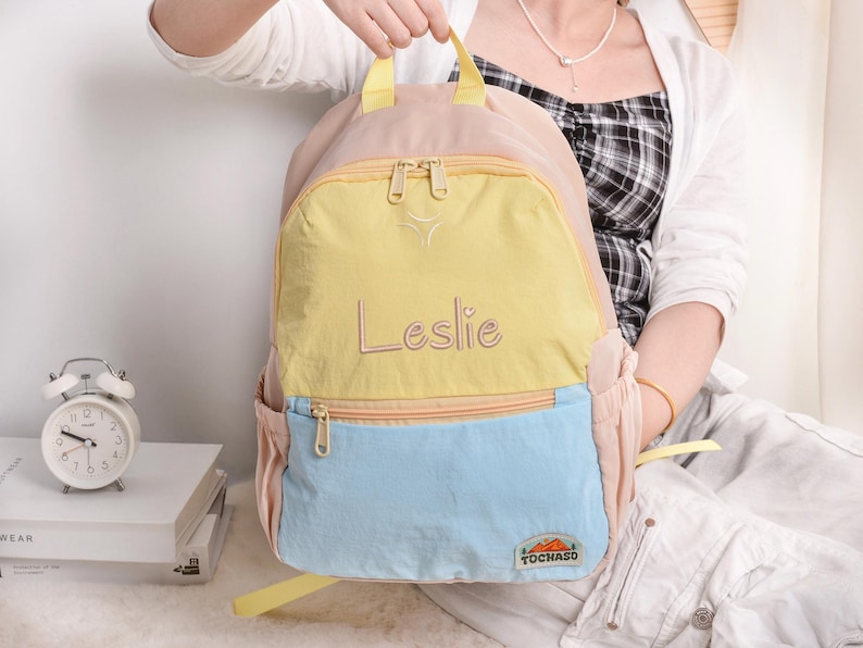 May include: A multi-colored backpack with a light pink body, a yellow front pocket, and a light blue bottom pocket. The name "Leslie" is embroidered in cursive on the yellow pocket. The backpack has a zipper closure and a small logo that says "TOCHASO".