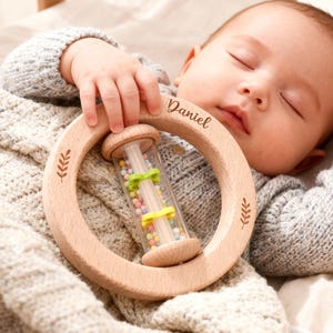 May include: A wooden baby rattle with the name "Daniel" engraved on it. The rattle features a clear tube filled with colorful beads and is held by a baby wearing a gray sweater. The rattle has leaf-like designs.