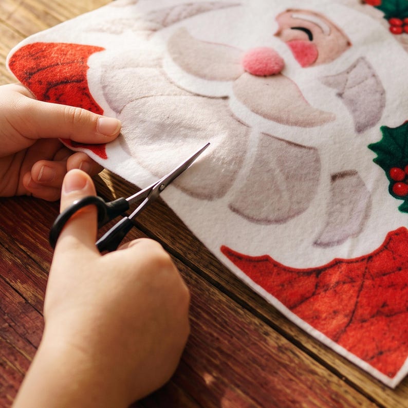 May include: A white felt craft sheet featuring a printed Santa Claus face with a pink nose, a red hat, and green holly leaves with red berries. The felt is being cut with black metal scissors, revealing the soft texture of the material.