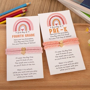 Puede incluir: Dos pulseras de amistad de color rosa con dijes de coraz&oacute;n dorados y una tarjeta con un dise&ntilde;o de arco&iacute;ris y el texto "first day of FOURTH GRADE" y "first day of PRE-K". La tarjeta incluye un mensaje sobre usar las pulseras en la escuela y pensar el uno en el otro.