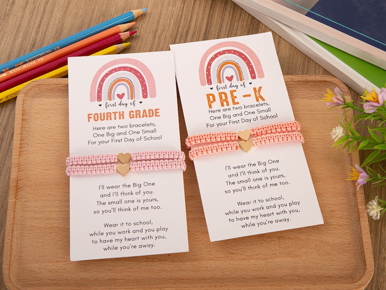 Puede incluir: Dos pulseras de amistad de color rosa con dijes de coraz&oacute;n dorados y una tarjeta con un dise&ntilde;o de arco&iacute;ris y el texto "first day of FOURTH GRADE" y "first day of PRE-K". La tarjeta incluye un mensaje sobre usar las pulseras en la escuela y pensar el uno en el otro.