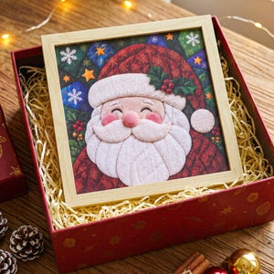 May include: A square wooden frame containing a quilted fabric art piece of Santa Claus. The design features a smiling Santa with a white beard, a red hat with holly berries, and a background of dark blue and green patches with white snowflakes and stars.