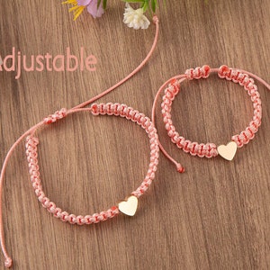 Puede incluir: Dos pulseras de amistad ajustables de color rosa con dijes de coraz&oacute;n de oro. Las pulseras est&aacute;n hechas de cord&oacute;n trenzado y est&aacute;n atadas con un nudo corredizo.