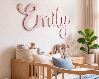 May include: A large, light pink wooden wall sign featuring the name Emily in a cursive script font. The letters are connected and mounted on a plain wall above a wooden crib, which is dressed with white and blue bedding and plush toys.