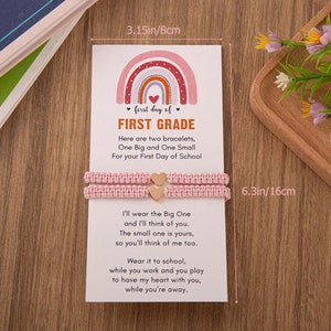 Puede incluir: Una tarjeta rosa y blanca con un dise&ntilde;o de arco&iacute;ris y el texto "first day of FIRST GRADE". La tarjeta est&aacute; acompa&ntilde;ada de dos pulseras de amistad rosas con dijes de coraz&oacute;n dorados. La tarjeta dice: "Aqu&iacute; hay dos pulseras, una grande y una peque&ntilde;a para tu primer d&iacute;a de escuela. Yo usar&eacute; la grande y pensar&eacute; en ti. La peque&ntilde;a es tuya, para que tambi&eacute;n pienses en m&iacute;. &Uacute;sala en la escuela, mientras trabajas y juegas para tener mi coraz&oacute;n contigo, mientras est&aacute;s lejos."