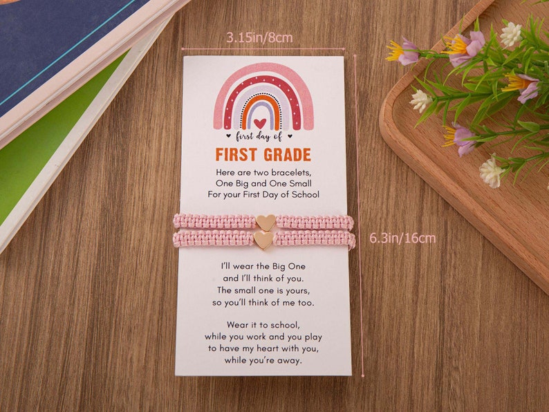 Puede incluir: Una tarjeta rosa y blanca con un dise&ntilde;o de arco&iacute;ris y el texto "first day of FIRST GRADE". La tarjeta est&aacute; acompa&ntilde;ada de dos pulseras de amistad rosas con dijes de coraz&oacute;n dorados. La tarjeta dice: "Aqu&iacute; hay dos pulseras, una grande y una peque&ntilde;a para tu primer d&iacute;a de escuela. Yo usar&eacute; la grande y pensar&eacute; en ti. La peque&ntilde;a es tuya, para que tambi&eacute;n pienses en m&iacute;. &Uacute;sala en la escuela, mientras trabajas y juegas para tener mi coraz&oacute;n contigo, mientras est&aacute;s lejos."