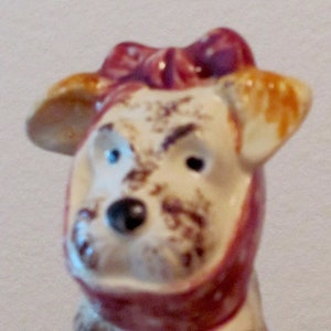 Può includere: Una statuetta di ceramica di un cane con un fiocco rosso e bianco. Il cane è seduto con le zampe ripiegate sotto di lui. La statuetta è dipinta con macchie marroni e bianche.