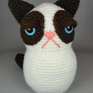 May include: A crocheted stuffed cat with brown ears, brown eyes, and a white body. The cat has a pink nose and a grumpy expression.