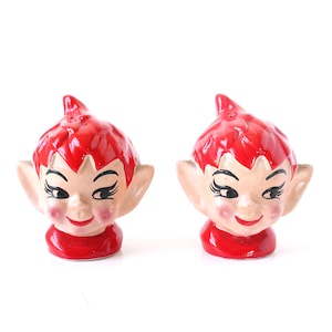 May include: A pair of vintage ceramic elf-shaped salt and pepper shakers. Each shaker features a red cap, rosy cheeks, and a smiling face. The shakers are painted with black eyelashes and a red collar.