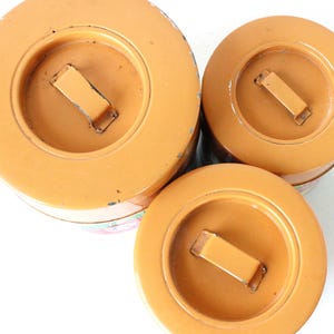 May include: Three round, orange metal canisters of varying sizes, each featuring a flat lid with a central rectangular handle. The canisters are arranged in a cluster, showing their top surfaces with visible signs of wear and paint chipping.
