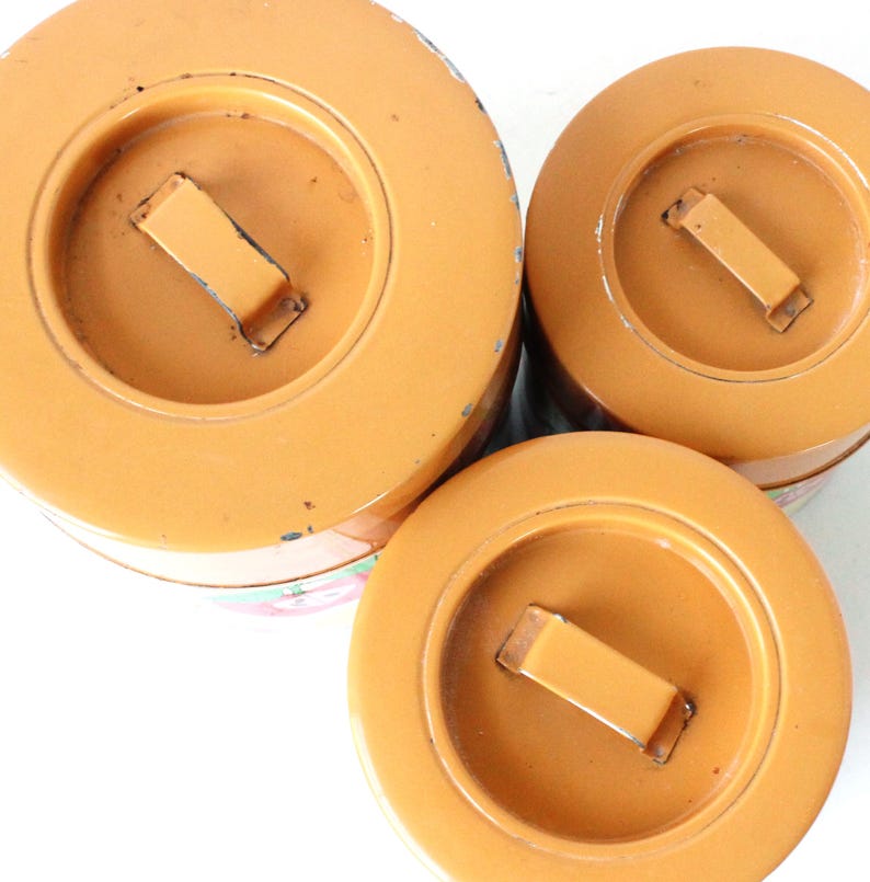 May include: Three round, orange metal canisters of varying sizes, each featuring a flat lid with a central rectangular handle. The canisters are arranged in a cluster, showing their top surfaces with visible signs of wear and paint chipping.