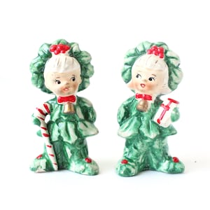 May include: Two vintage Christmas figurines. Each figurine is a smiling girl with a green dress and a holly wreath headdress. One holds a candy cane, the other a gift. Both have red bows and bells.