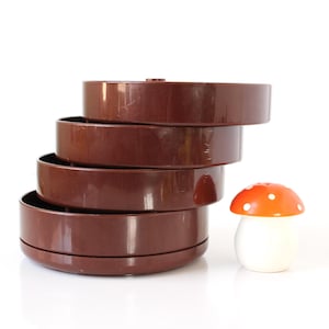 May include: A brown plastic tiered storage container with four circular rotating compartments stacked vertically. Beside the storage unit sits a small, white, mushroom-shaped shaker with an orange cap featuring white polka dots.