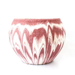 Könnte beinhalten: Ein runder, dekorativer Pflanztopf mit Marmormuster. Der Pflanztopf hat ein wirbelndes Muster aus weißen und staubrosafarbenen Farben. Der Topf besteht aus einem strukturierten Material und steht vor einem weißen Hintergrund.