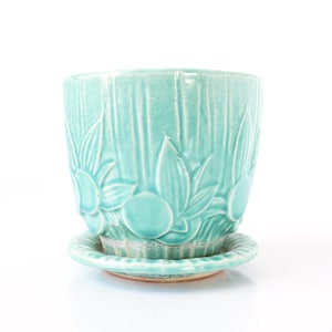 Pode incluir: Um vaso de flores de cerâmica turquesa com um pires a condizer. O vaso apresenta um design texturizado e canelado com padrões florais e de folhas em relevo. O vaso foi concebido para conter plantas e flores.