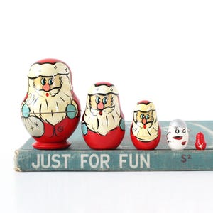 May include: A set of five nesting dolls, each painted with a Santa Claus design. The dolls are red, white, and black, with varying sizes. The dolls are arranged on a blue book with the text "JUST FOR FUN".
