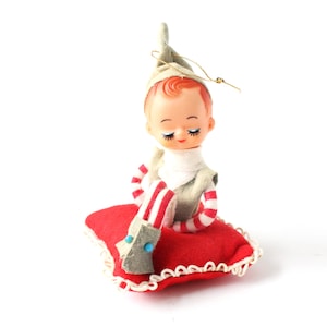Puede incluir: Adorno navideño vintage con una figura de muñeca con los ojos cerrados, que lleva un gorro y un chaleco verdes. La figura está sentada sobre una almohada roja con ribete blanco y tiene piernas a rayas rojas y blancas. Una cuerda dorada está unida al gorro.
