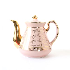 Könnte beinhalten: Eine Vintage-Teekanne in Rosa mit goldenen Akzenten. Die Teekanne hat einen goldenen Griff, Ausguss und Deckelknauf. Der Korpus ist mit einem goldenen geometrischen Muster und floralen Akzenten verziert. Ein klassisches Stück für Teepartys.