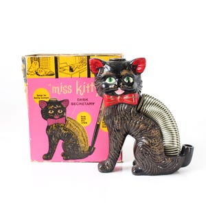 May include: A black ceramic cat desk organizer with green eyes, red ears, and a red bow tie. A metal coil spring extends from the cat's back to hold papers. The cat is positioned in front of its original pink and yellow box labeled Miss Kitty Desk Secretary.