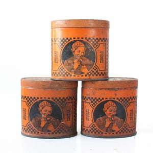 May include: Three cylindrical orange metal tins stacked in a pyramid formation. Each tin features a black circular graphic of a person wearing a bonnet and a checkered pattern, with the text MARQUE DEPOSEE and MODELE DEPOSE printed on the sides.