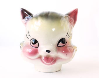 May include: A vintage ceramic cat head planter with a smiling expression. The cat has pink cheeks, black eyes, and a pink tongue. The ears are pink and the top of the head is striped with green and gray.