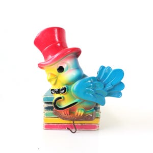 May include: A colorful bird-shaped wall hook with a red top hat, blue wings, and a black bow tie. The bird is perched on a stack of colorful blocks. The hook is made of a glossy material.