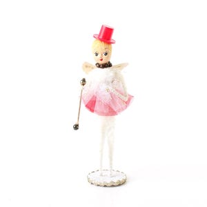 May include: A decorative figurine featuring a doll with blonde hair wearing a bright red top hat. The doll has white wings, a pink tulle skirt, and long white pipe cleaner legs. It holds a thin gold-toned wand with a small dark bead at the end.