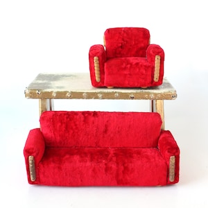 May include: A miniature red velvet sofa and armchair set. The furniture sits on a small, rustic table with a distressed, off-white top and gold-colored accents. The sofa has two cushions and the armchair has armrests.