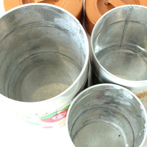 May include: Three cylindrical metal canisters of varying sizes with open tops and silver-toned interiors. The exterior surfaces feature a white base with colorful fruit illustrations, while the lids are solid orange with small metal handles.