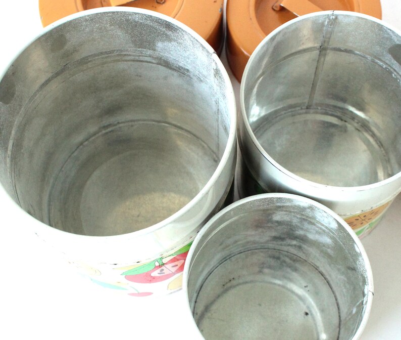 May include: Three cylindrical metal canisters of varying sizes with open tops and silver-toned interiors. The exterior surfaces feature a white base with colorful fruit illustrations, while the lids are solid orange with small metal handles.