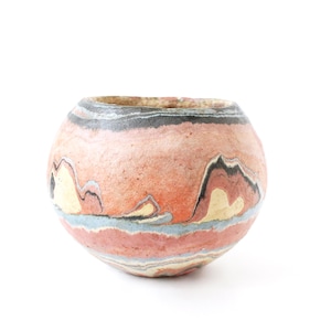 Könnte beinhalten: Ein runder, handgefertigter Keramiktopf mit einem einzigartigen, geschichteten Design. Der Topf weist Farbbänder in Pfirsich, Schwarz, Grau und Blau auf, wodurch ein Marmoreffekt entsteht. Das Design ähnelt einer Landschaft. Der Topf ist ein dekorativer Gegenstand.