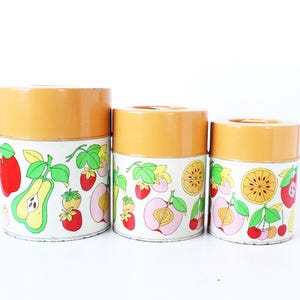 May include: A set of three cylindrical metal canisters with orange lids and white bodies featuring a colorful fruit pattern. The design includes illustrations of red apples, green pears, strawberries, cherries, peaches, and sliced citrus fruits.