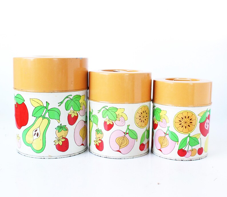 May include: A set of three cylindrical metal canisters with orange lids and white bodies featuring a colorful fruit pattern. The design includes illustrations of red apples, green pears, strawberries, cherries, peaches, and sliced citrus fruits.
