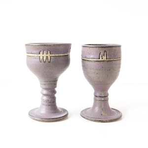 Peut inclure: Deux gobelets en céramique faits à la main, de couleur lavande douce. Chaque gobelet présente une bande décorative avec un motif doré près du bord. Le pied et la base sont également lavande, créant un aspect artistique et cohérent.