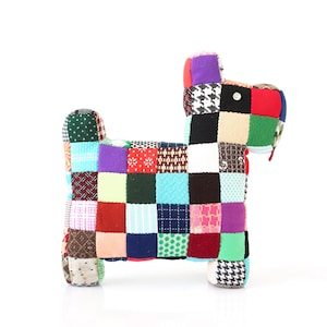 Könnte beinhalten: Ein hundeförmiges Stofftier aus einem Patchwork aus bunten Stoffquadraten. Der Hund hat ein Knopfauge und einen kleinen roten Faden als Schwanz. Der Patchwork umfasst Blumen-, Tupfen- und geometrische Muster.